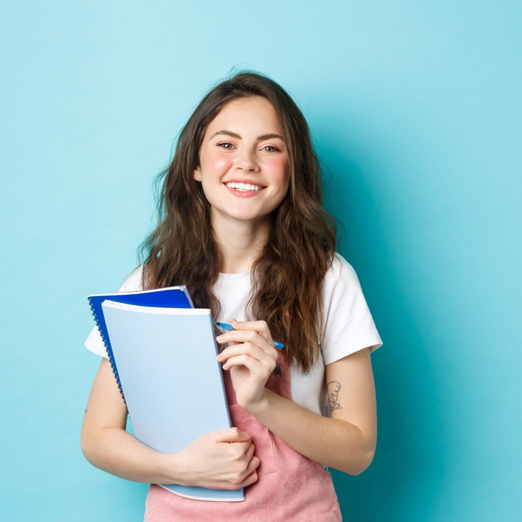 happy young female student holding notebooks from courses and smiling at camera, standing in spring clothes against blue background